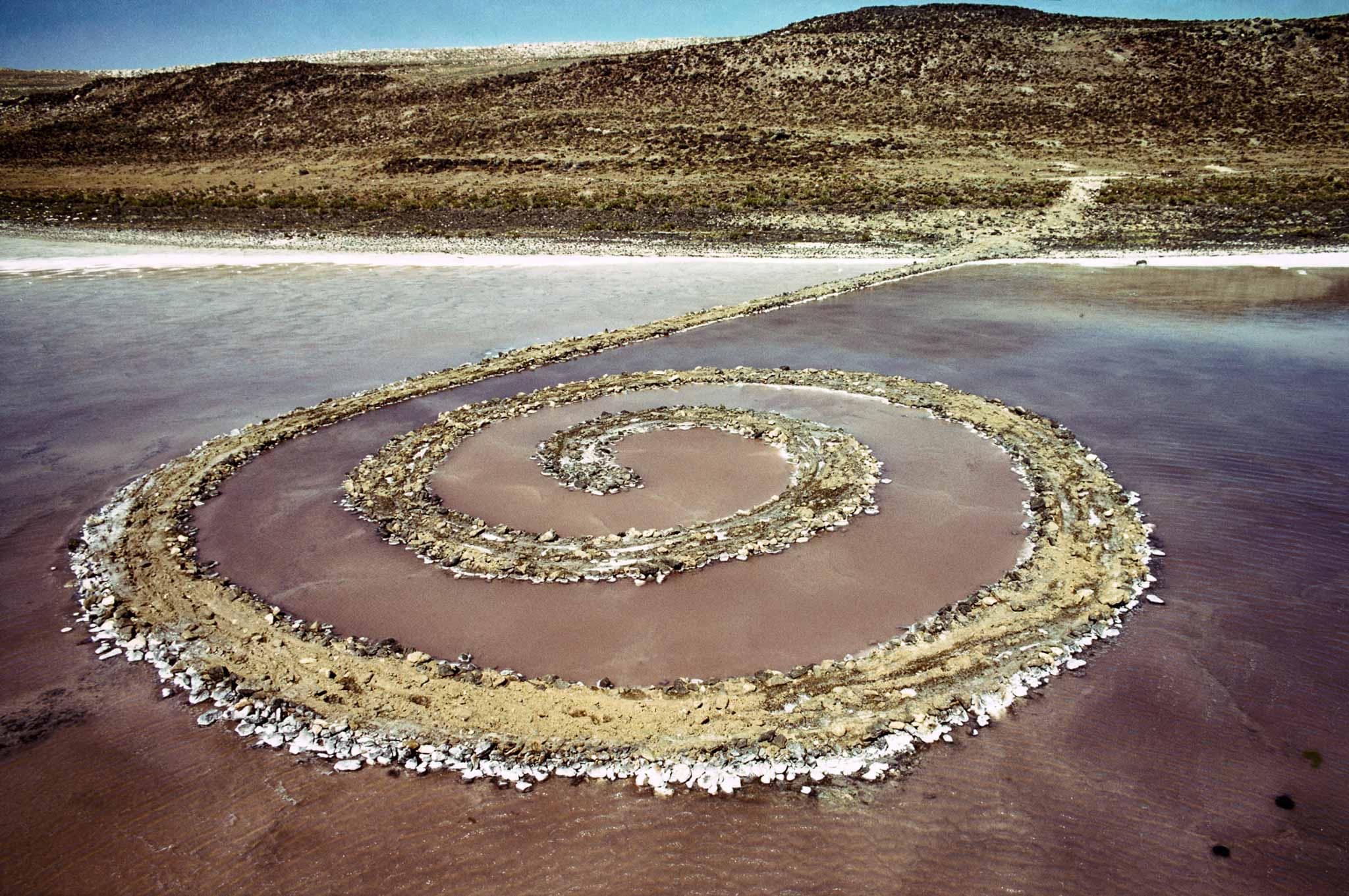 Robert Smithson, Spiral Jetty [Σπειροειδής αποβάθρα], 1970, Great Salt Lake, Γιούτα, ΗΠΑ. Ο Smithson κατασκεύασε τη μνημειακή σπειροειδή κατασκευή χρησιμοποιώντας περίπου 6.650 τόνους βράχων, χώματος και άλλων φυσικών υλικών, δημιουργώντας μια προεξοχή μήκους περίπου 460 μέτρων και πλάτους περίπου 4,5 μέτρων που εκτείνεται από την όχθη προς τη λίμνη. Η Spiral Jetty αποτελεί εμβληματικό έργο της γεωτέχνης, καθώς η μορφή της συνδέεται άμεσα με το φυσικό τοπίο και υπόκειται στις μεταβολές της στάθμης του νερού, αναδεικνύοντας τη σχέση ανάμεσα στην καλλιτεχνική παρέμβαση, τη γεωλογική διαδικασία και τη χρονικότητα.