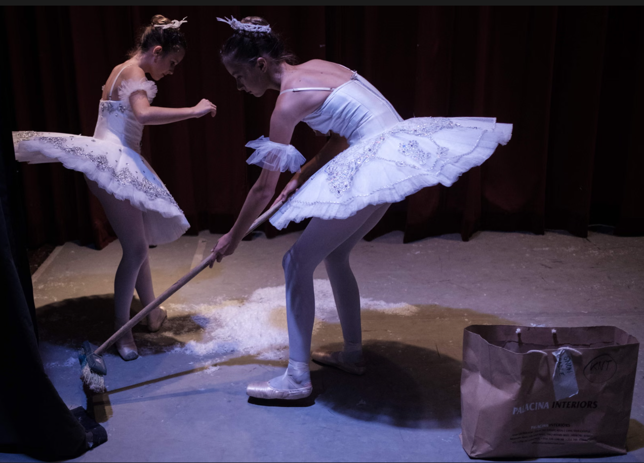 Δύο νεαρές χορεύτριες του [[#Dance Centre Kenya [Κέντρο Χορού Κένυας]#]] καθαρίζουν τις τεχνητές νιφάδες χιονιού από τη σκηνή κατά το διάλειμμα παράστασης του Καρυοθραύστη [The Nutcracker]. Φορώντας ακόμη τα λευκά τους τουτού, αναλαμβάνουν εργασίες σκηνικής συντήρησης που κανονικά παραμένουν αόρατες, αποκαλύπτοντας το διάλειμμα ως χρόνο πρακτικής, παρασκηνιακής μέριμνας πίσω από τη μαγεία του θεάματος. Φωτογραφία: Yasuyoshi Chiba/AFP/Getty Images.